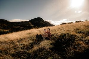 People sitting on a grassy hilltop during golden hour with tall grass swaying in the wind and mountains in the background.