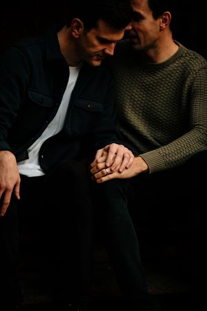 Two people hold hands in a dark setting wearing dark pants and a green knit sweater.