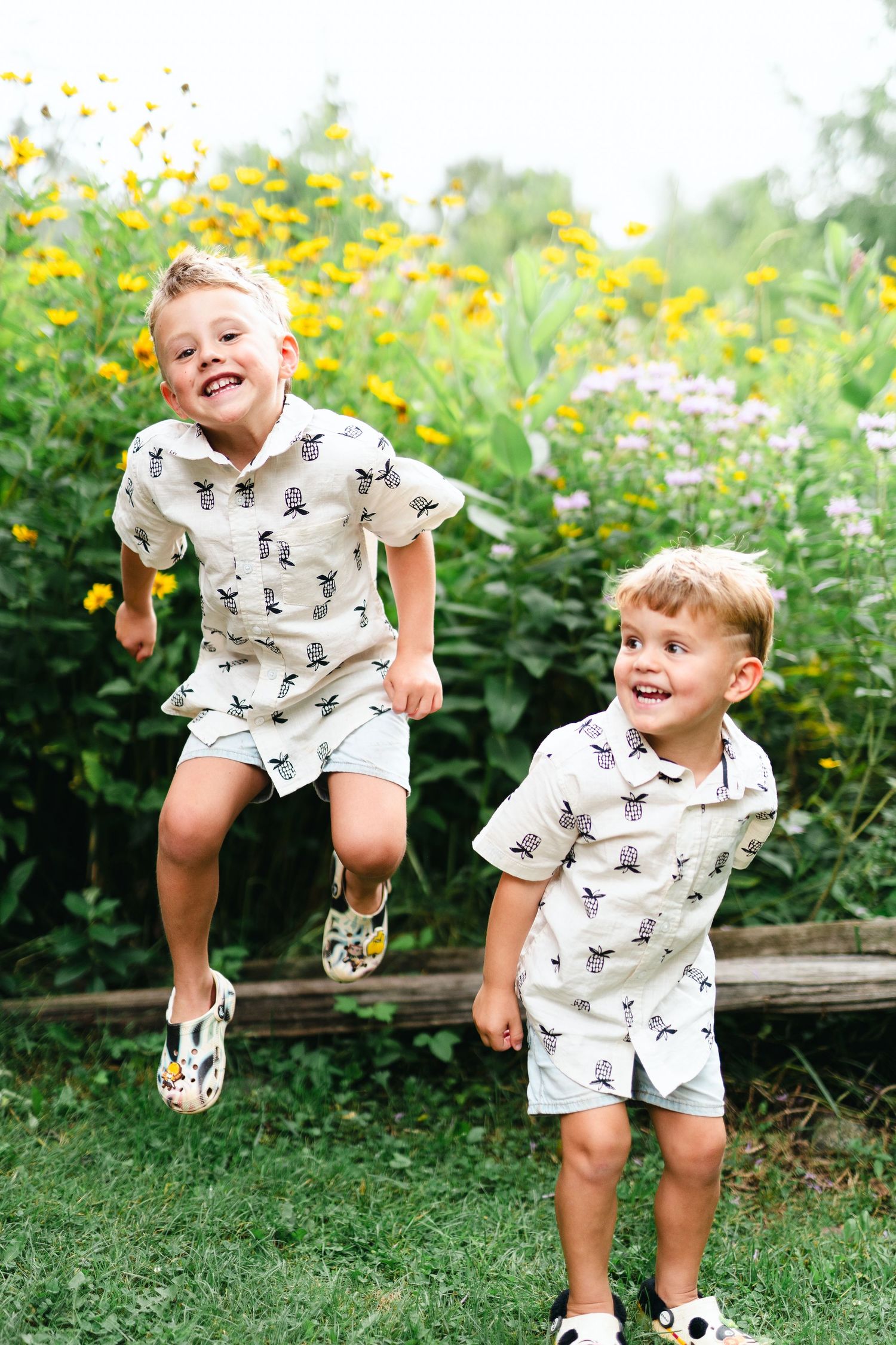 Two children in matching dinosaur print outfits play and jump together in a garden with yellow flowers.