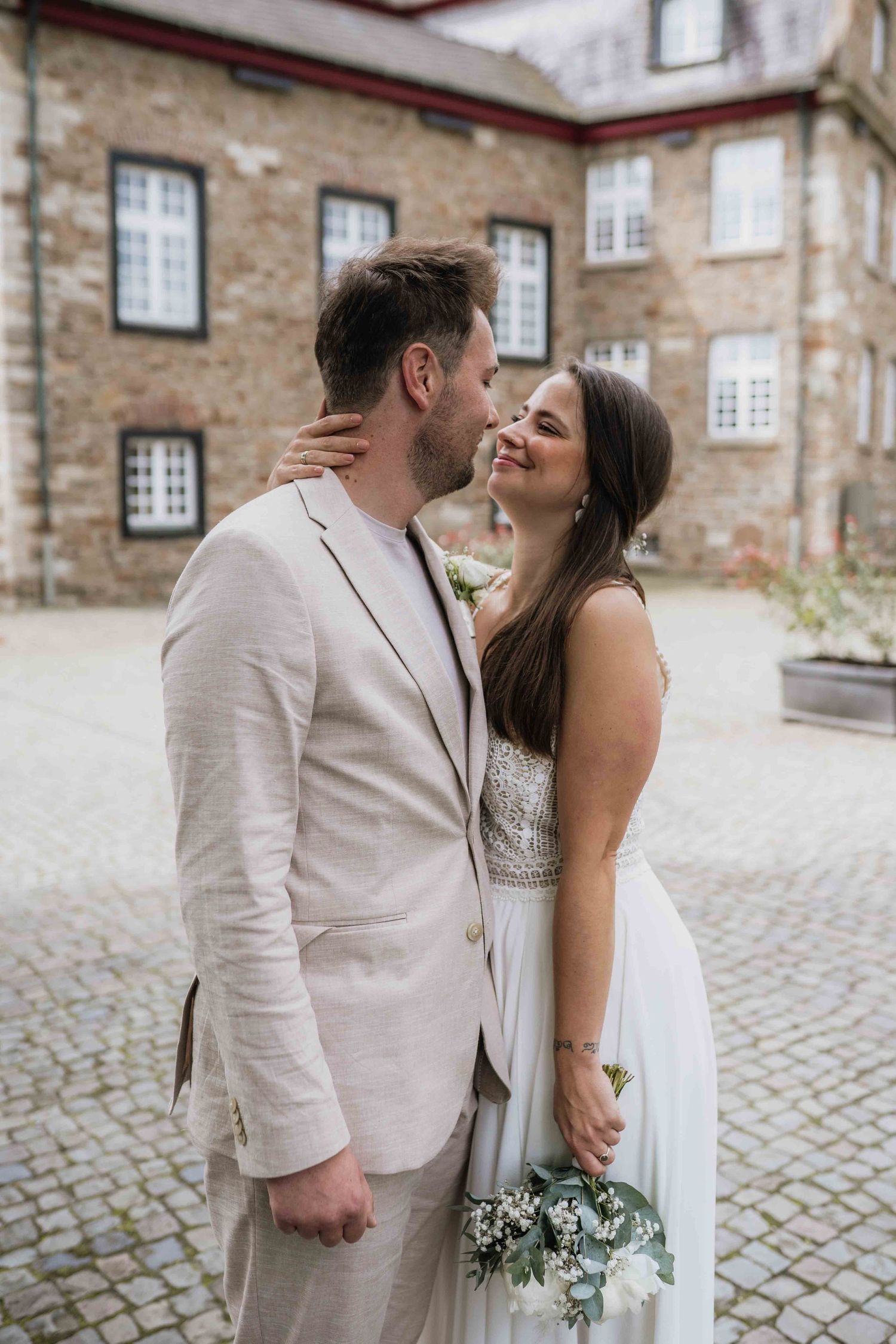 Romantic couple in light-colored wedding attire embracing in courtyard.