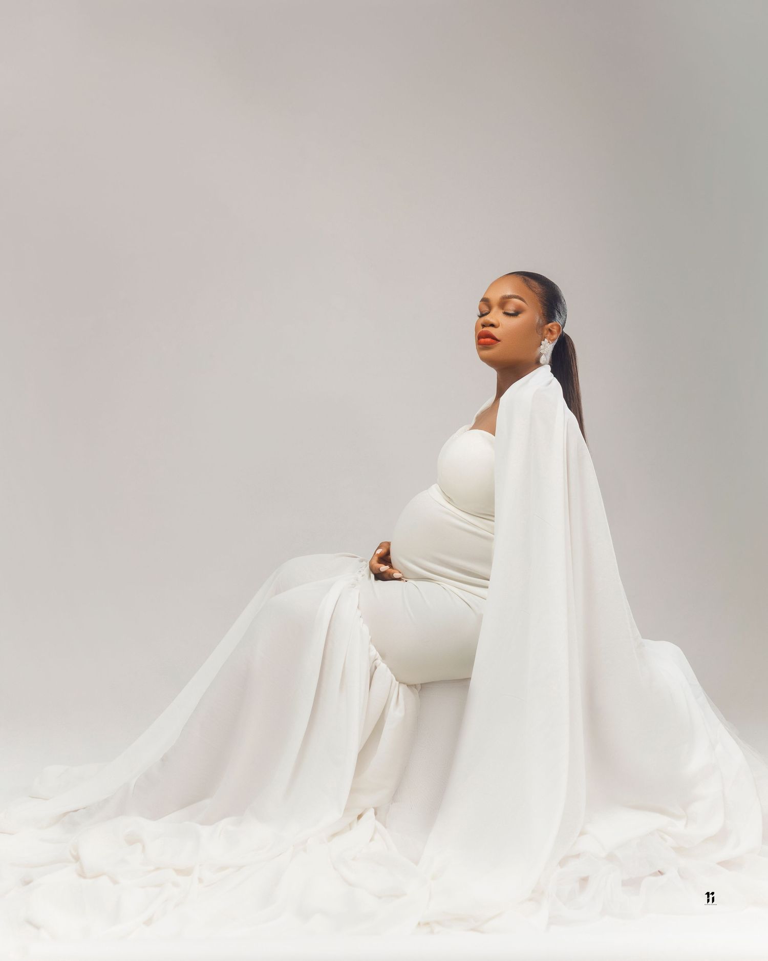 Artistic maternity portrait in white flowing dress against light background.