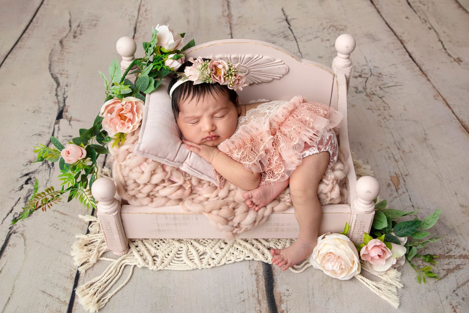 Sleeping baby in pink lace outfit on miniature bed with roses and greenery decoration.