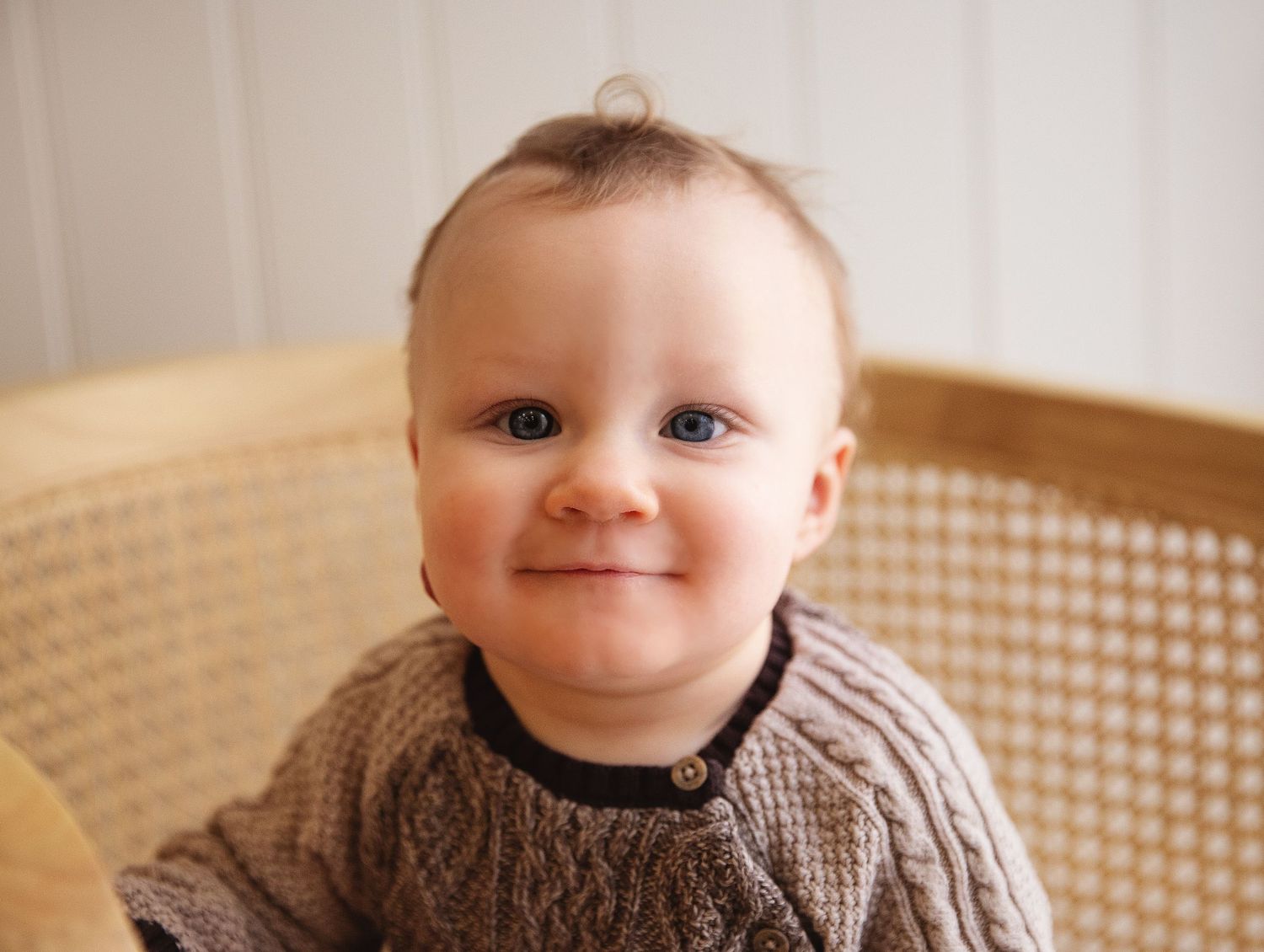 A small child wearing a cozy knit sweater smiles while sitting in a cane-backed chair.