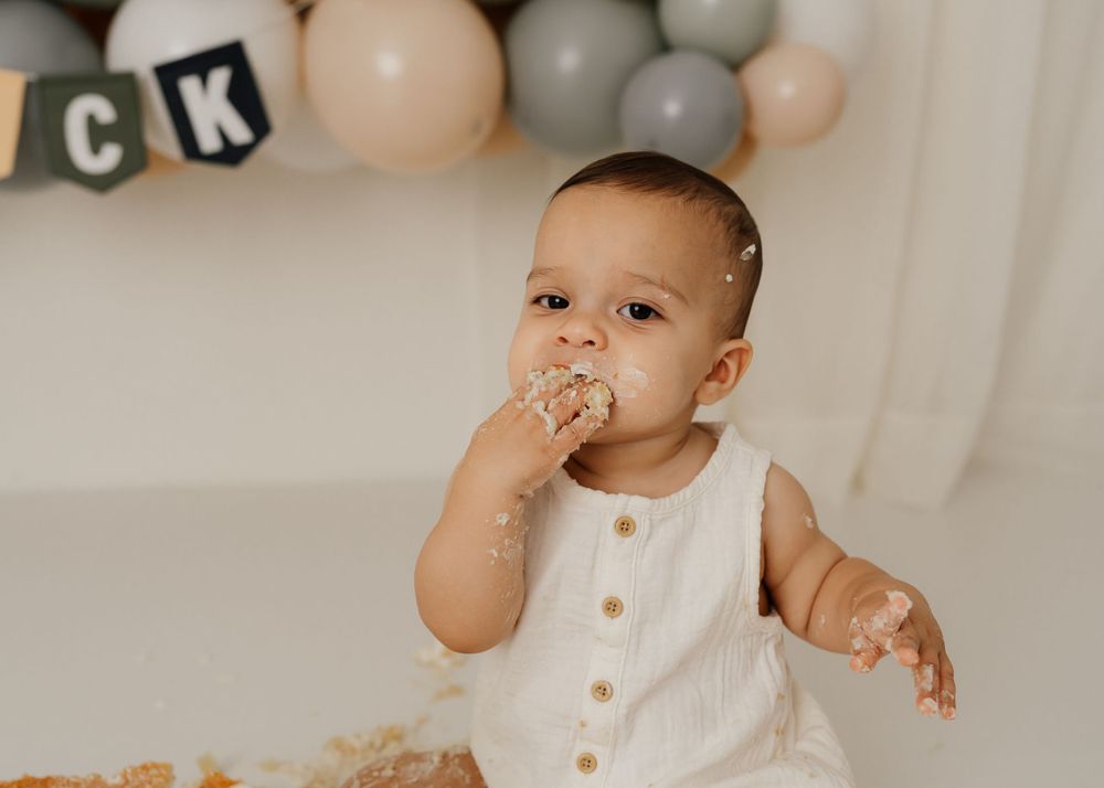 A baby tastes birthday cake against a backdrop of neutral colored balloons.