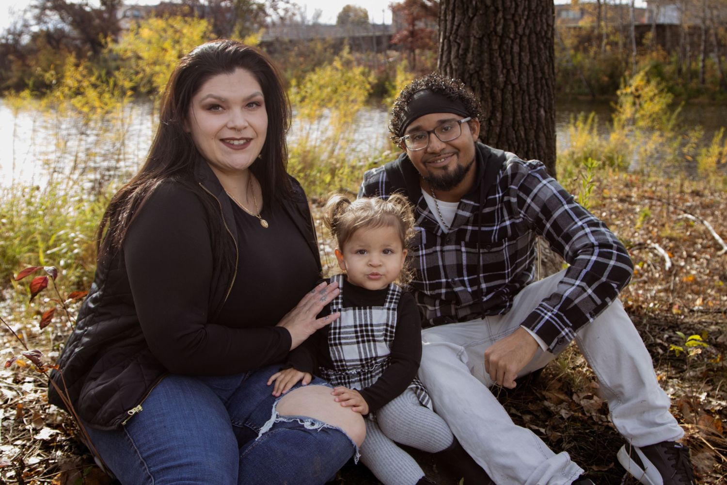 A family sits together outdoors near a tree and water during an autumn photoshoot.