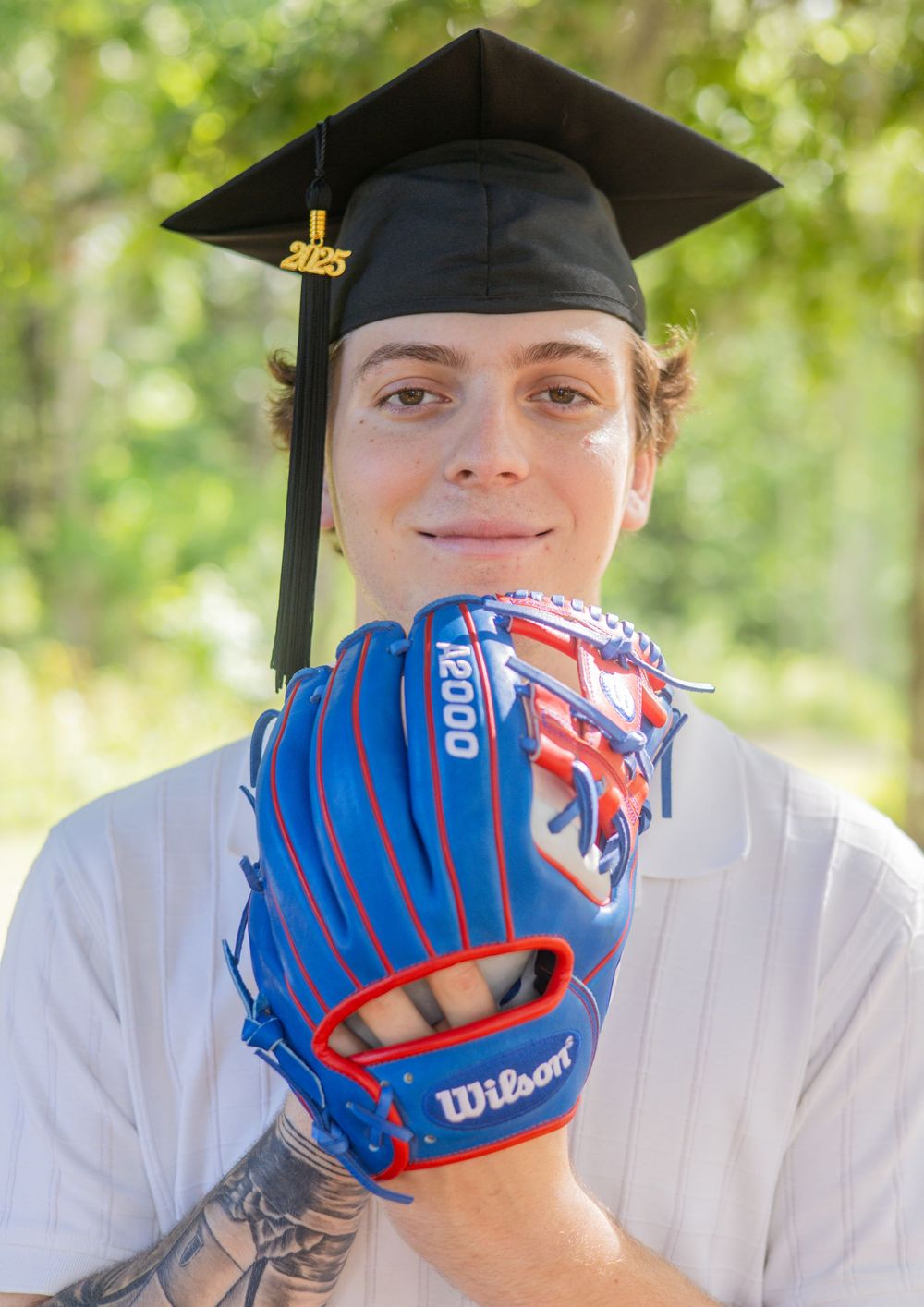 A graduation portrait featuring a blue baseball glove and black cap and gown against a natural outdoor background.