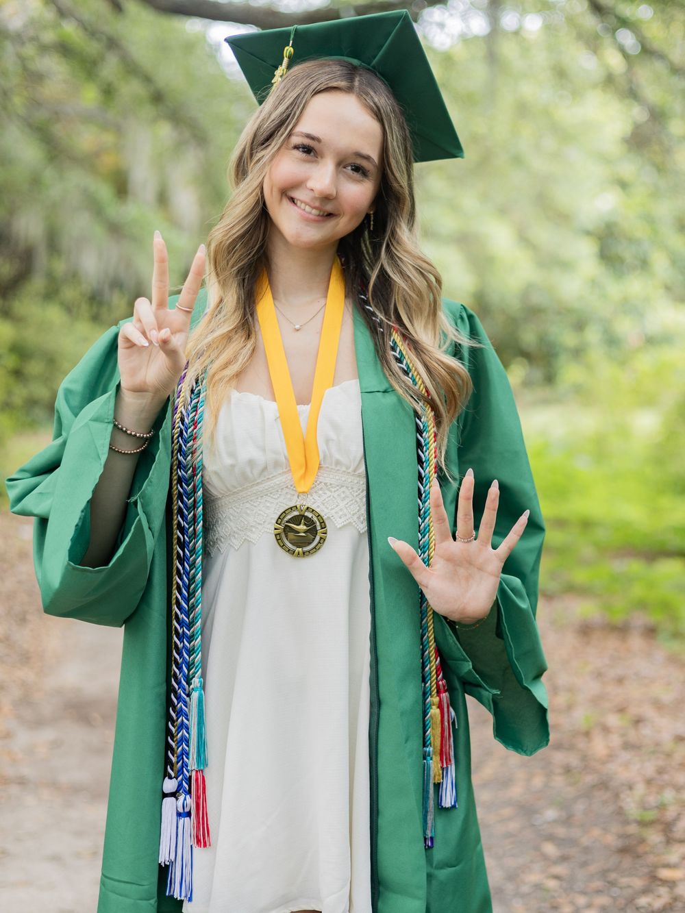Graduate in green cap and gown wearing gold medal and honor cords makes peace sign and waves at camera outdoors.