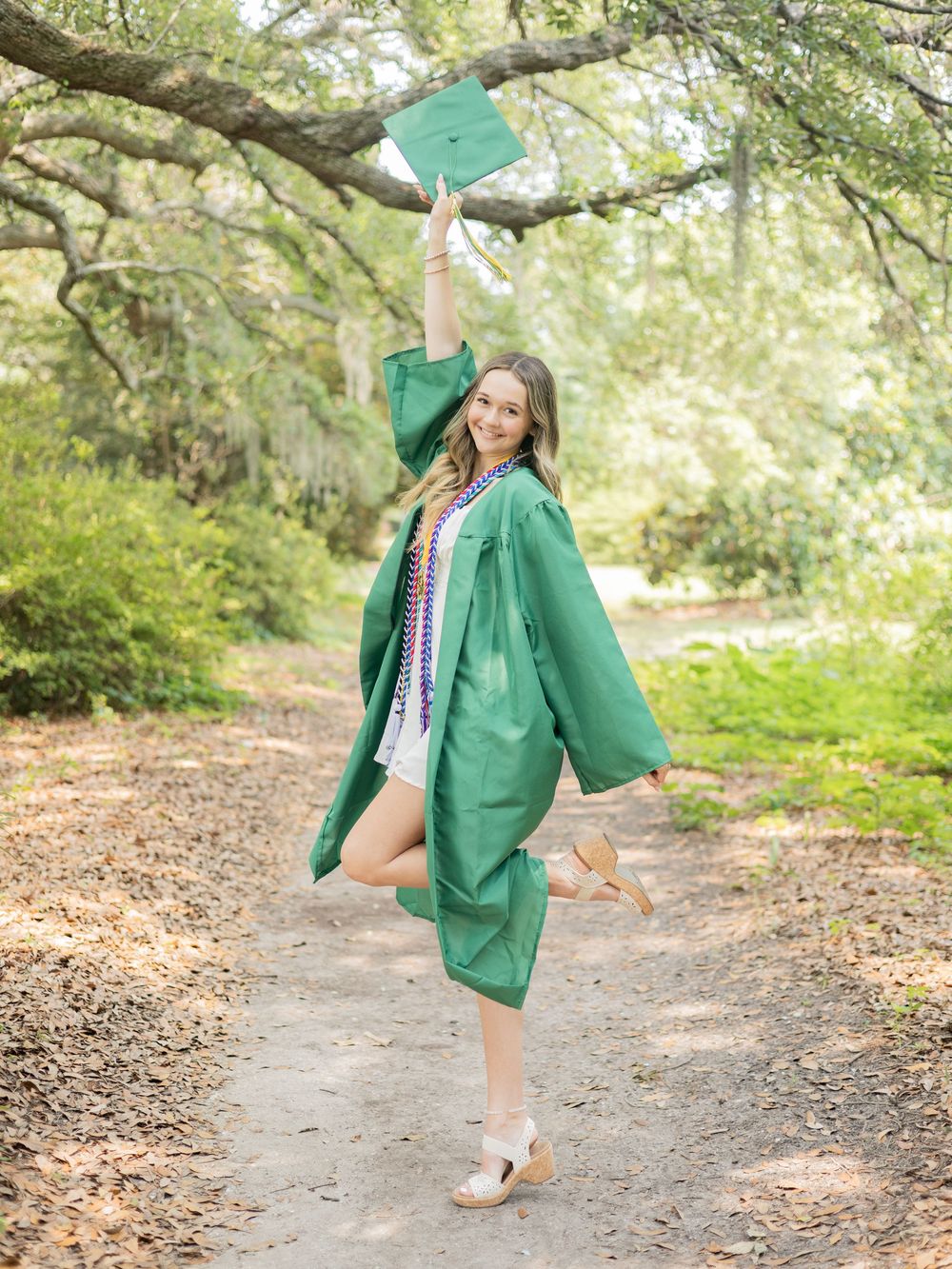 Graduate in emerald green cap and gown poses joyfully on a nature path while tossing graduation cap in the air.
