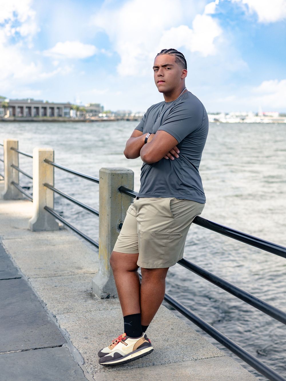 Person in gray shirt and khaki shorts stands contemplatively by waterfront railing on cloudy day.