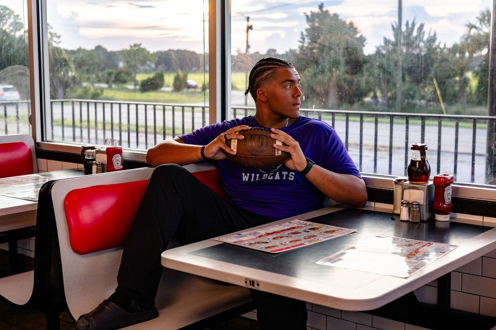 Someone sits at a table in a casual dining restaurant with large windows overlooking water and palm trees outside.