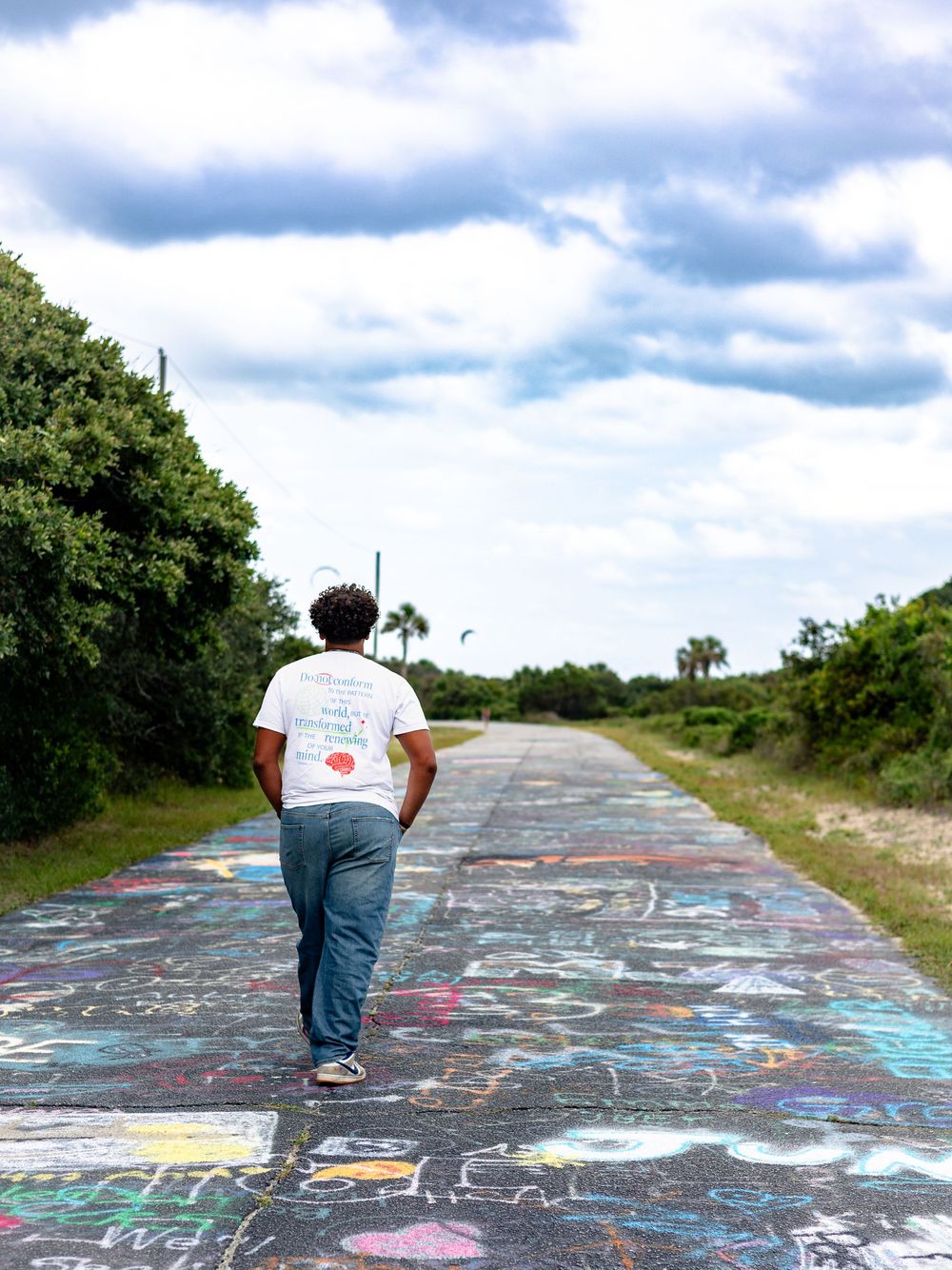 Someone walking on a colorful graffiti-covered path through a natural area with palm trees and cloudy sky.