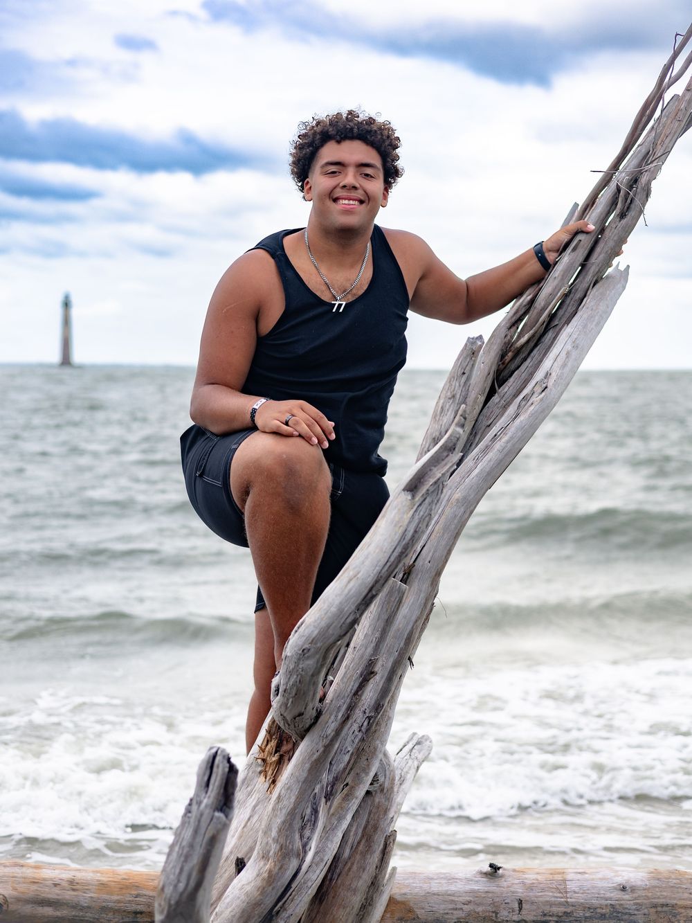 A person in a black tank top sits and smiles on a piece of driftwood at the beach with waves in the background.