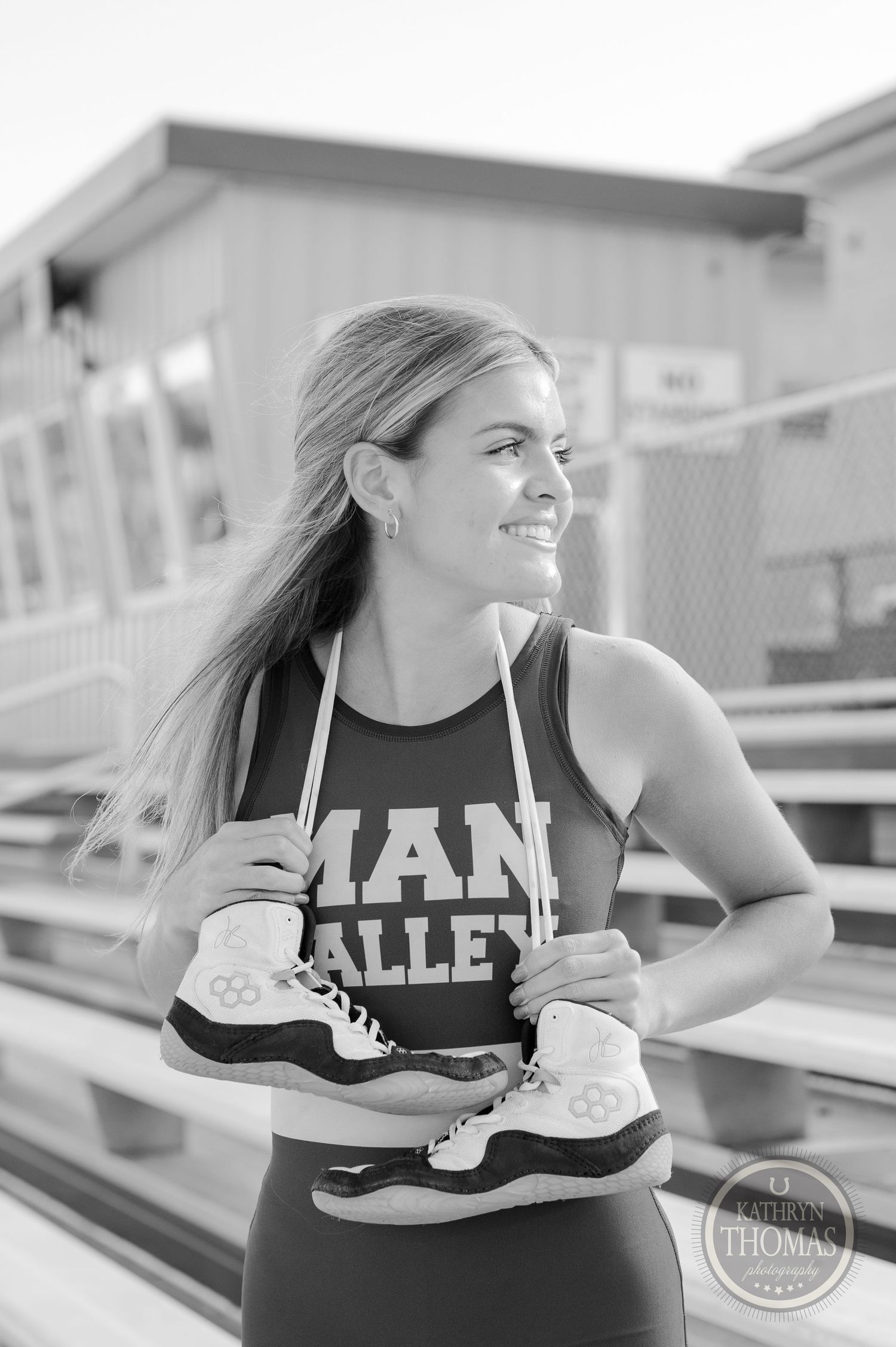 Black and white portrait of athlete holding sneakers while posing on stadium bleachers.