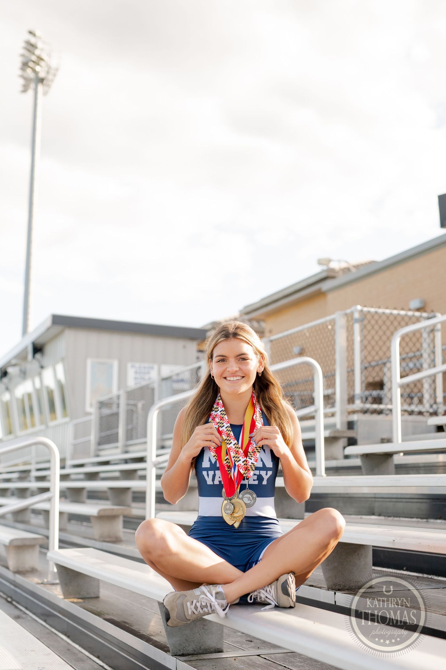 Student athlete displaying medals while sitting cross-legged on stadium bleachers with lights in background.