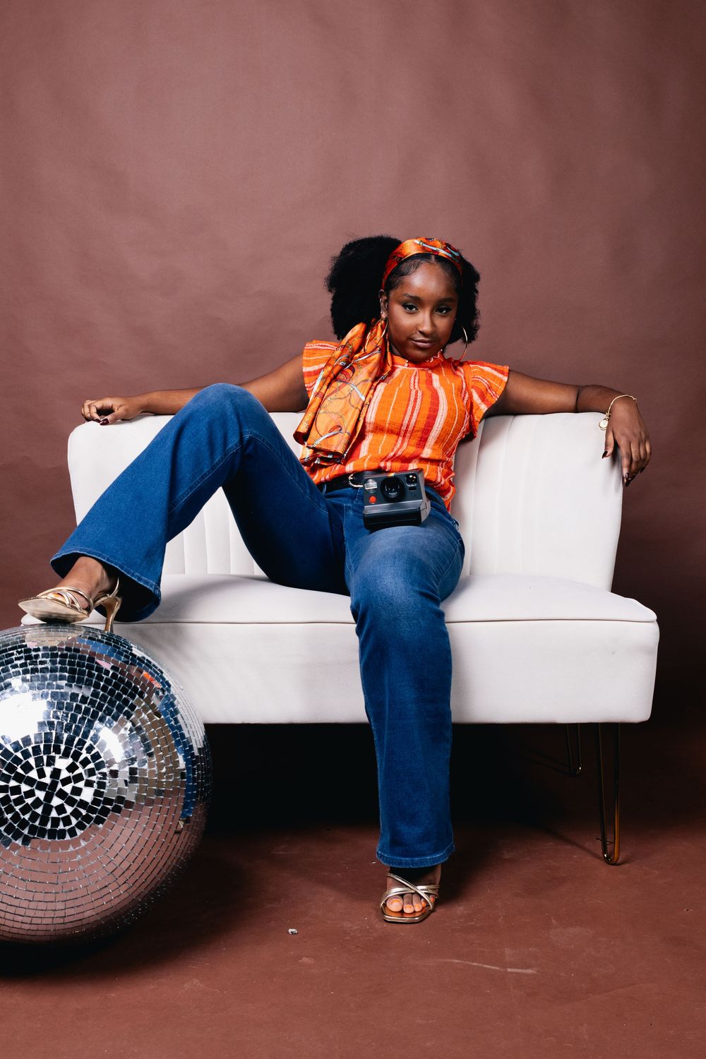 A young Black woman in orange top and flare jeans poses on a white chair beside a disco ball in a studio.