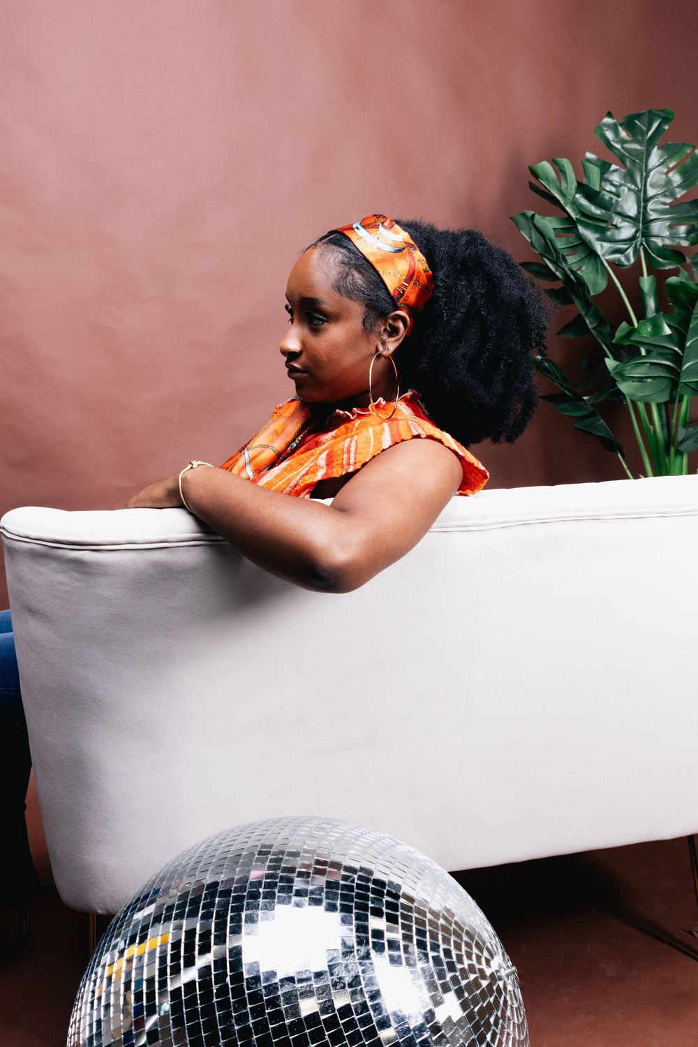 A young Black woman in an orange outfit and headband leans over a white sofa near a disco ball and tropical plant.