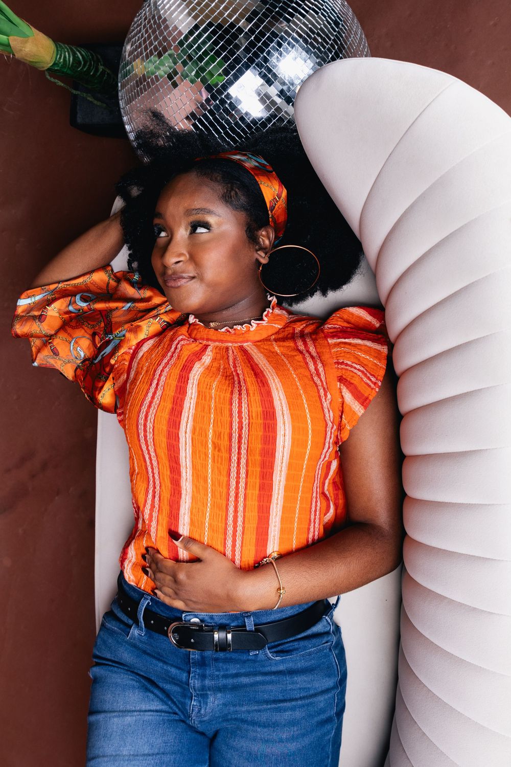 Young Black woman in orange striped top and jeans lying beside white couch with disco ball overhead.