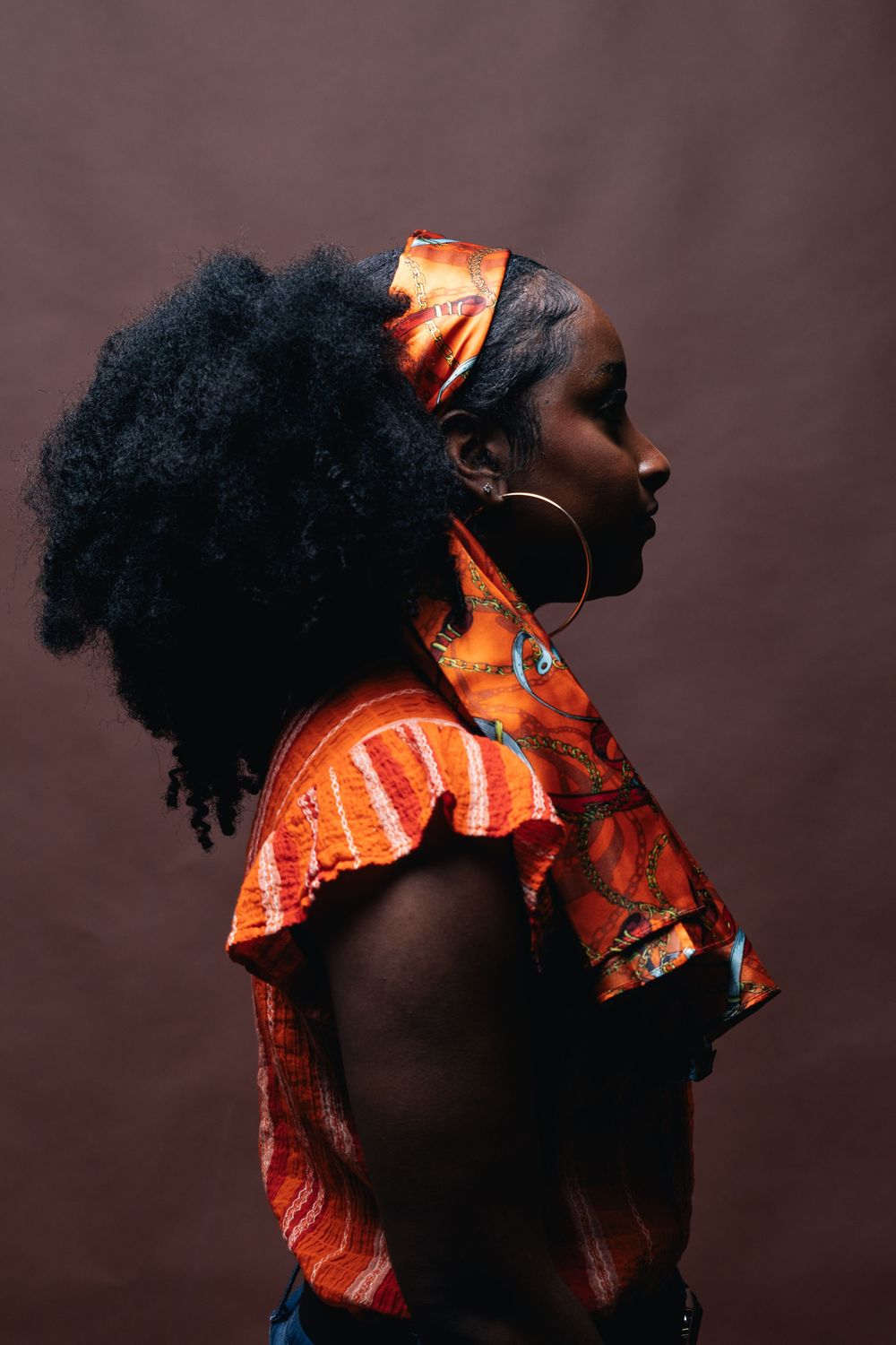 Profile view of a Black woman in an orange African print top and hoop earrings, with natural afro hair against a dark background.