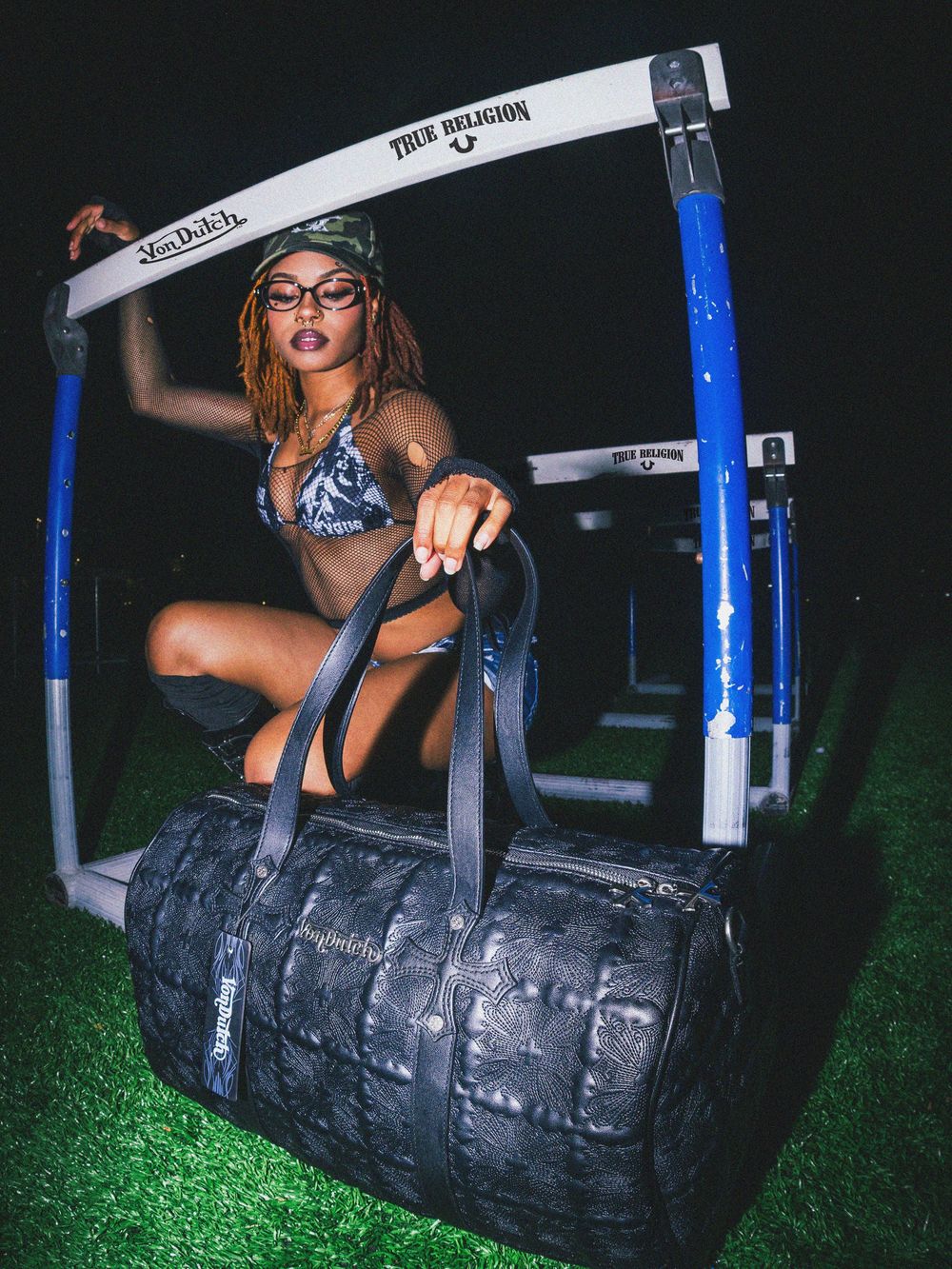 A person in athletic wear poses with a large tire and exercise equipment at night on green-lit turf.