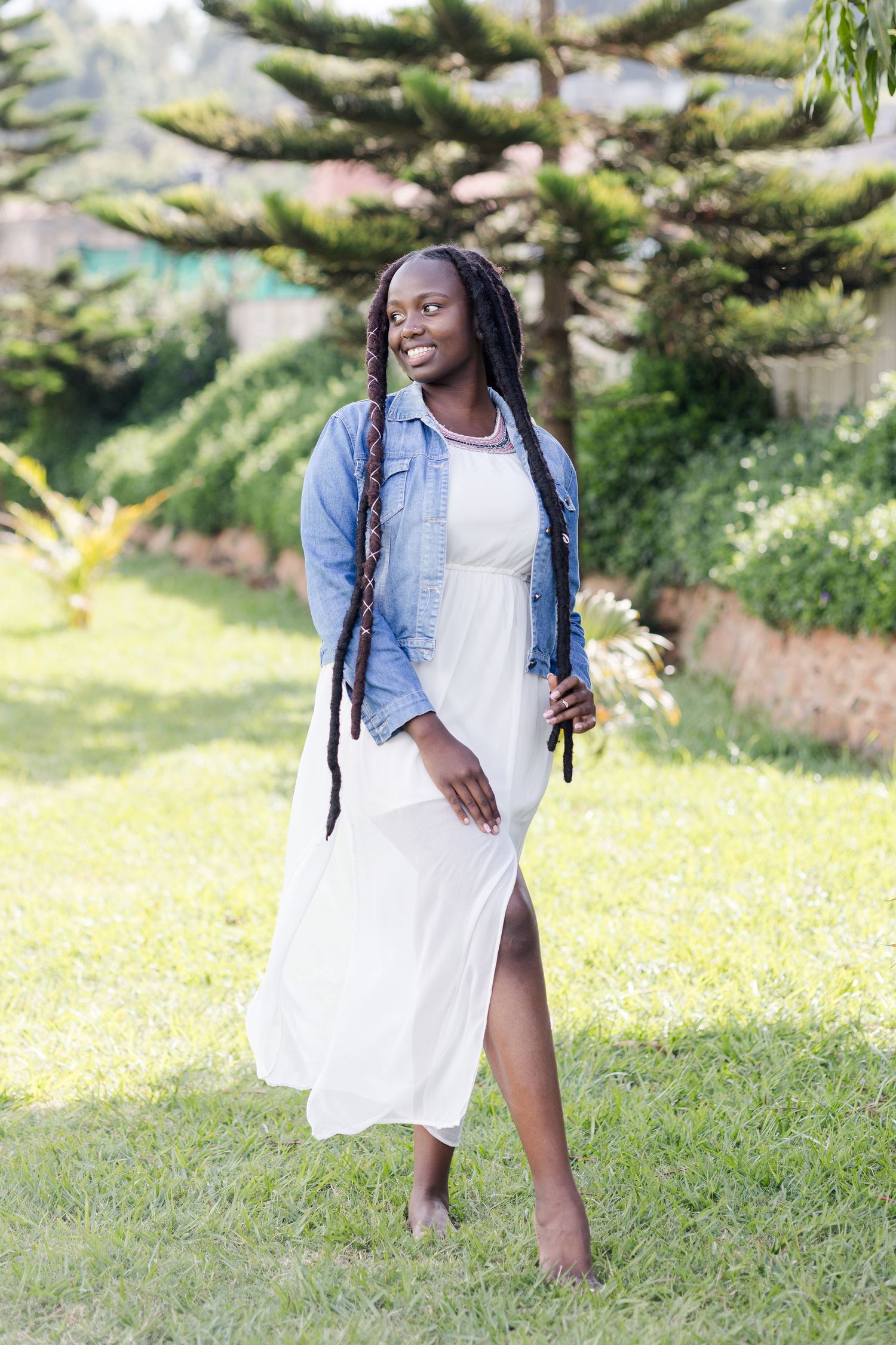 Full-length shot of a lady in white dress and blue jacket. She has thick locs. Standing with legs crossed. Ikigu George Photography.
