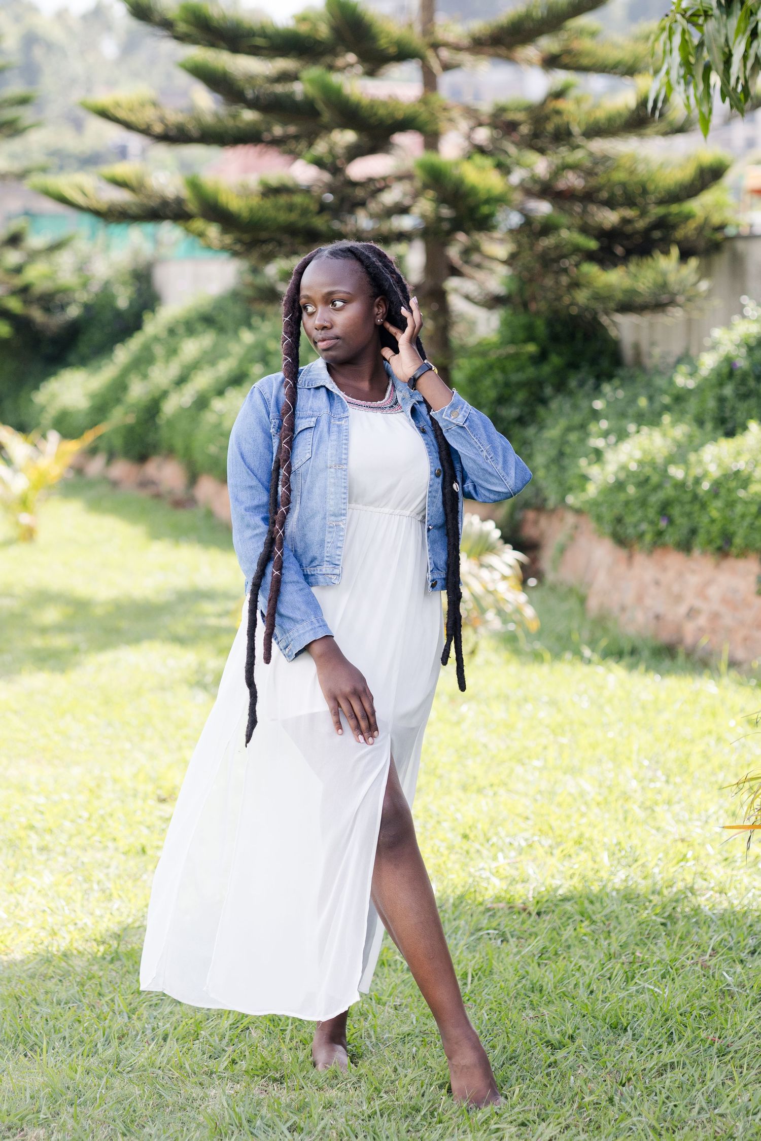 Lady in white dress with a slit and a denim jacket. Legs crossed. Left hand up to her ear. Ikigu George Photography.