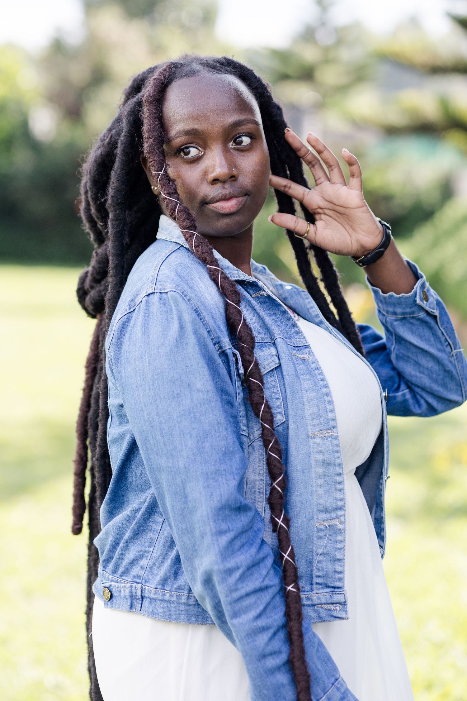 Mid-length portrait of lady in white dress & denim jacket. She has thick locs, and her left hand up to her ear. Ikigu George Photography.