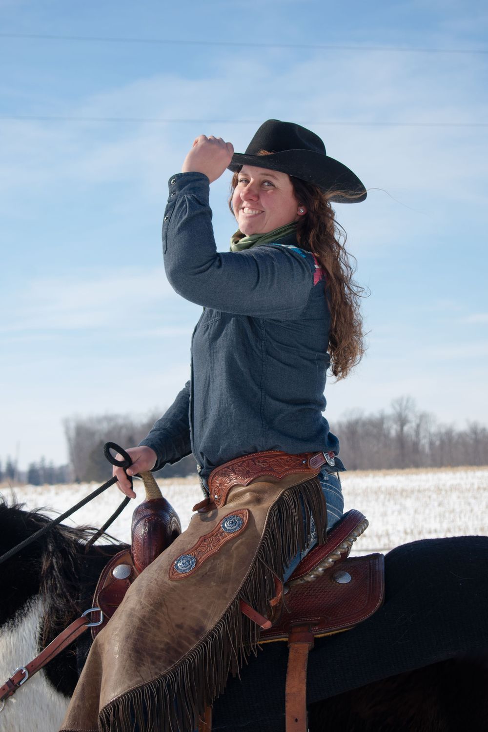 Cowgirl holding hat brim smiling over shoulder while riding her horse in a Wisconsin winter scene