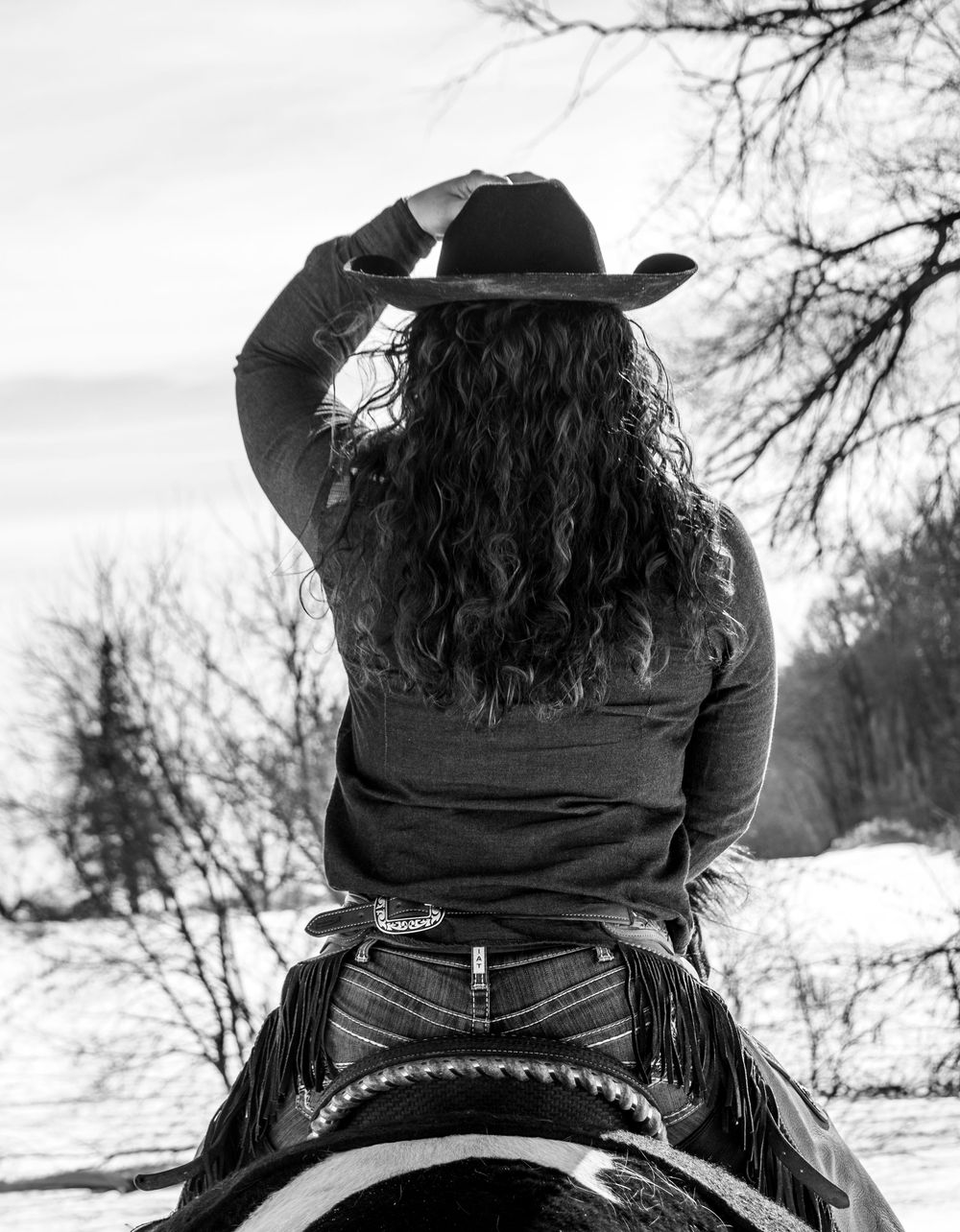 Black and white photo of cowgirl riding horse facing away while holding her hand on the top of her hat in a Wisconsin winter scene
