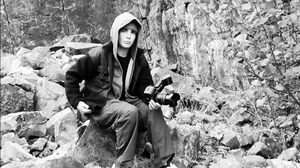 A person in a hoodie sits on rocky terrain in black and white photography.