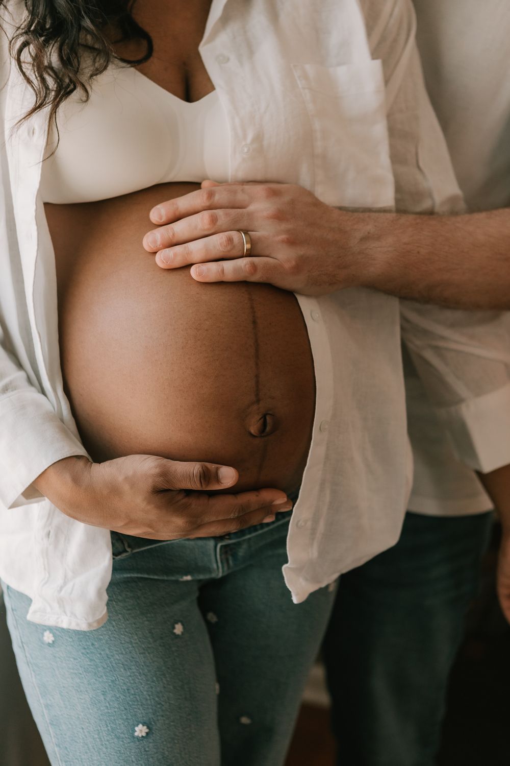 A close-up of a pregnant belly with hands tenderly cradling the bump, wearing a white shirt and denim jeans.