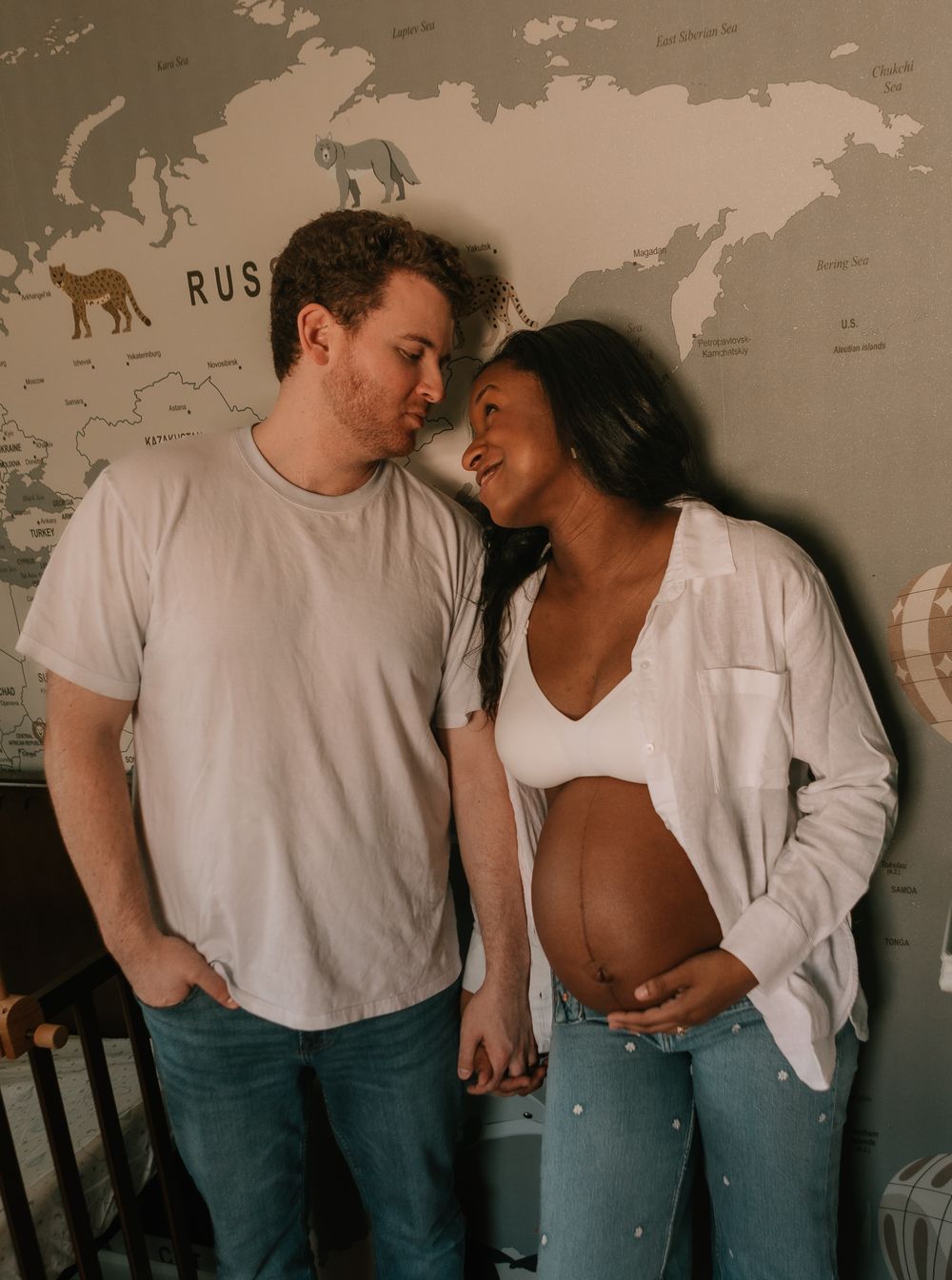 A couple stands intimately in front of a world map wall, with the pregnant person wearing a white crop top and open shirt.