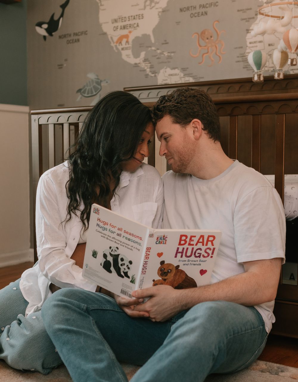A couple sits together on the floor in a nursery with a children's book titled 'Bear Hugs' against a world map wall.