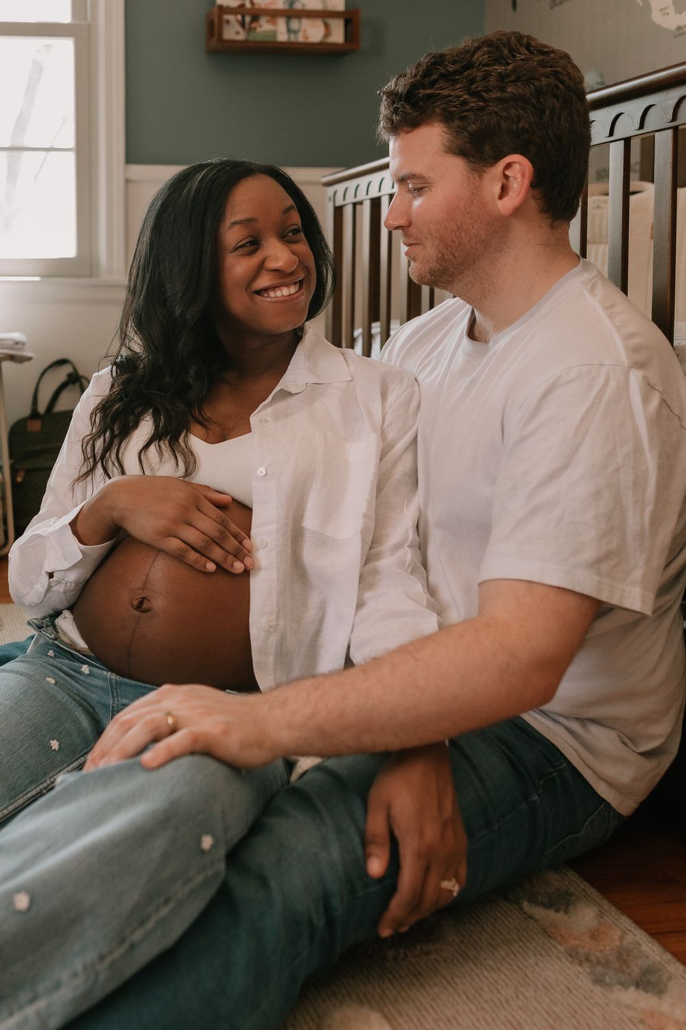 Expectant couple sits on floor sharing intimate moment, with white button-down shirt revealing pregnant belly.