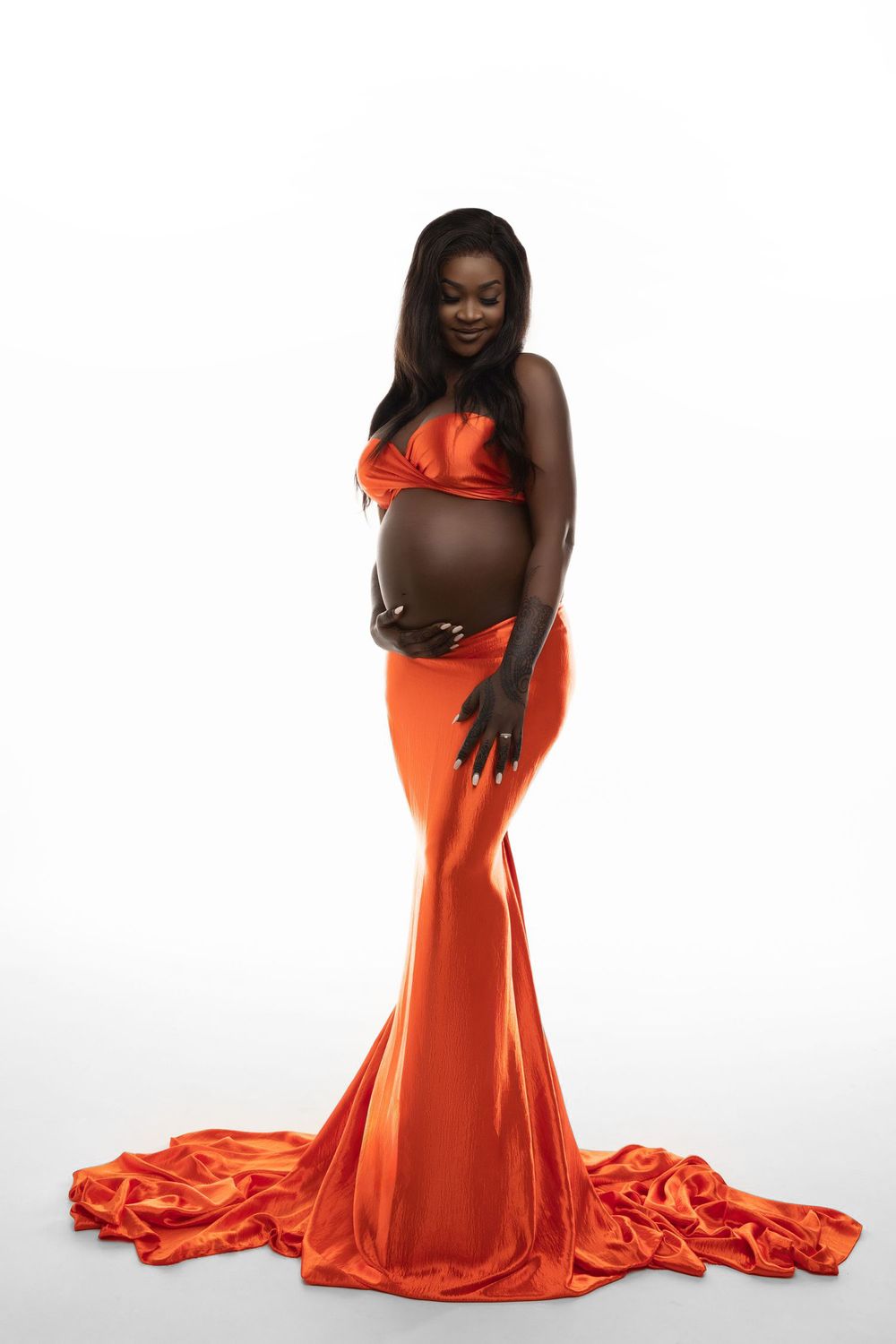 A pregnant woman poses in a flowing orange mermaid-style gown with a long train against a white backdrop.