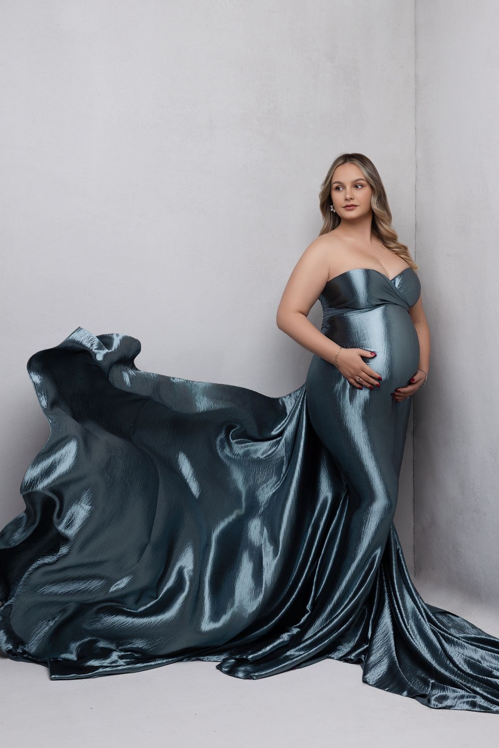 Elegant maternity photoshoot featuring a flowing teal silk gown with dramatic train against a minimalist grey backdrop.