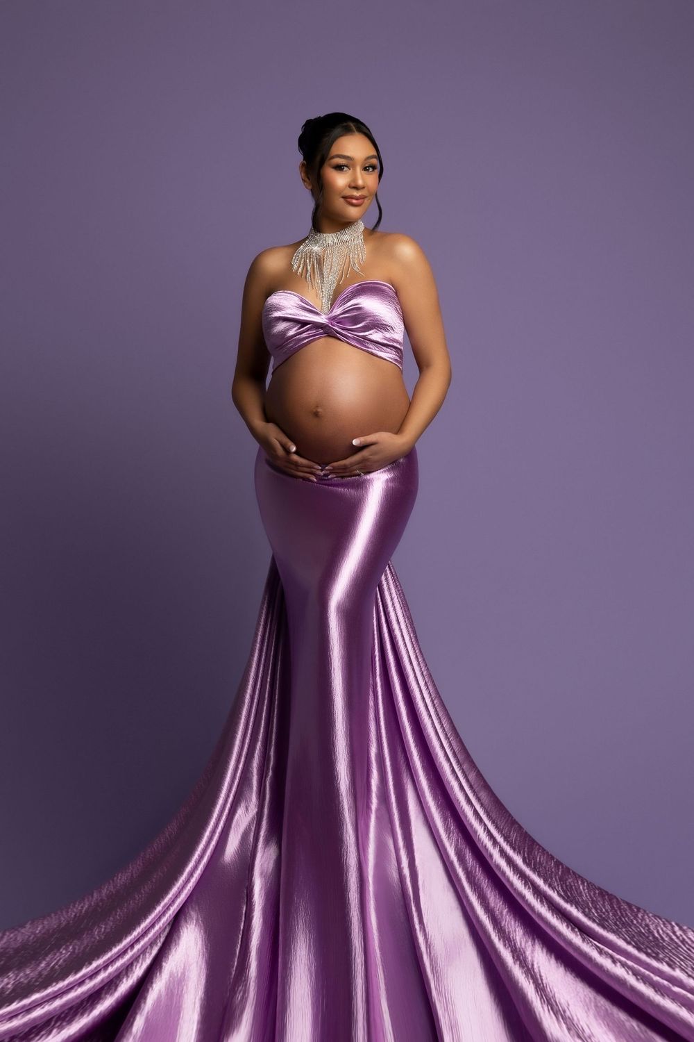 Expectant mother poses in flowing purple metallic maternity gown against purple backdrop in elegant studio portrait.