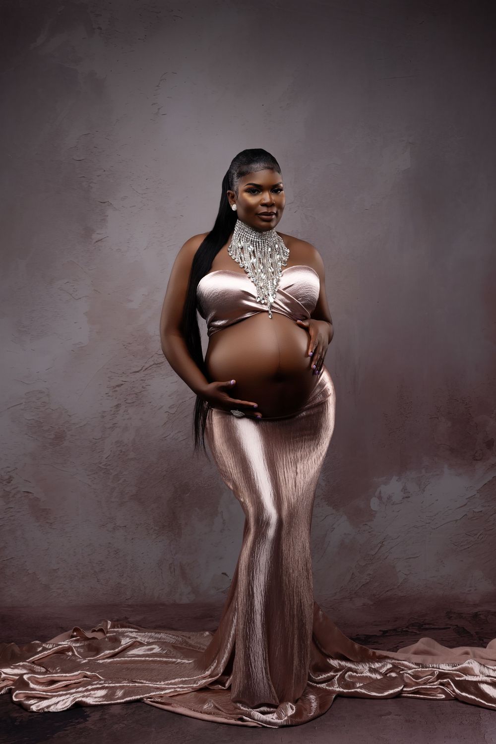 An elegant maternity portrait in rose gold dress with lace detail against dark background showcases a pregnant silhouette.