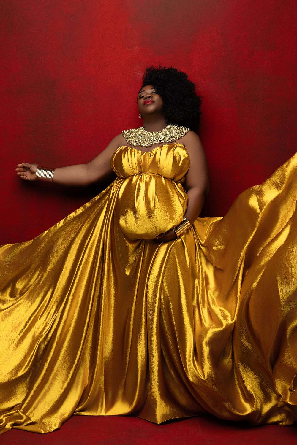 A person in a flowing golden satin gown poses dramatically against a deep red backdrop.