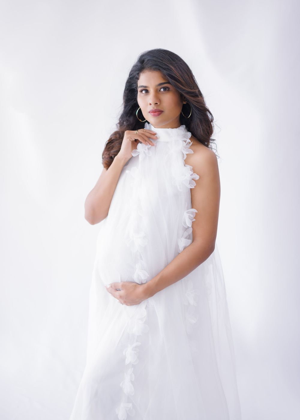 Elegant white high-neck maternity gown with ruffled details against light backdrop.