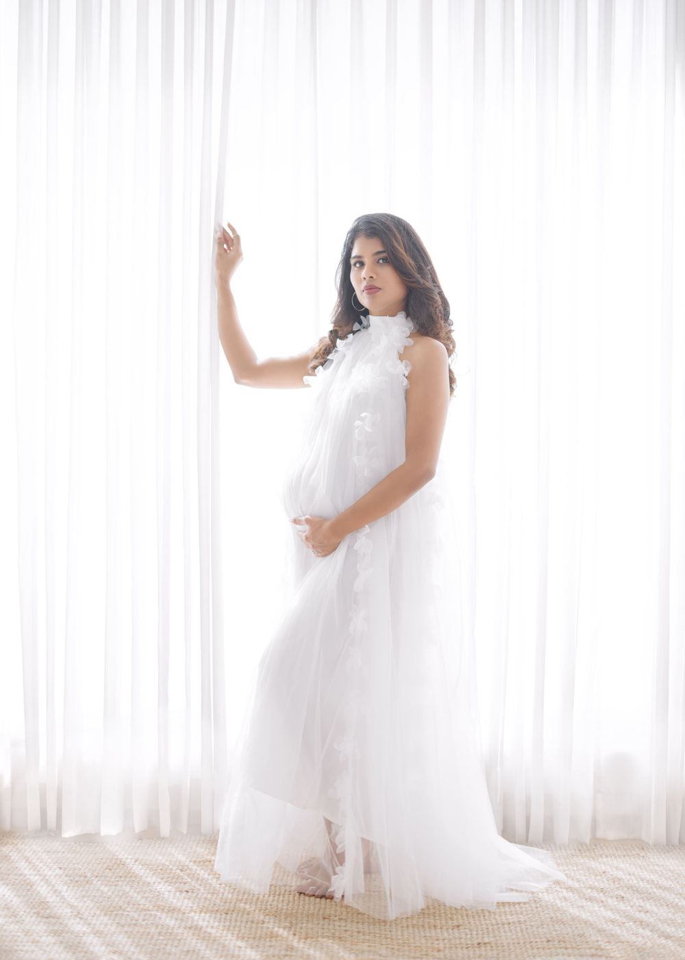 Dreamy backlit maternity portrait in flowing white dress against sheer curtained window.