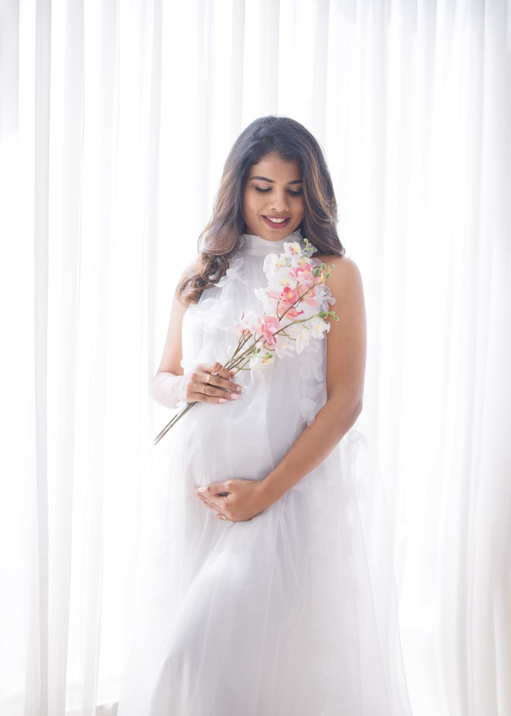 A maternity photo featuring a white lace dress and delicate pink cherry blossom branch against white curtains.