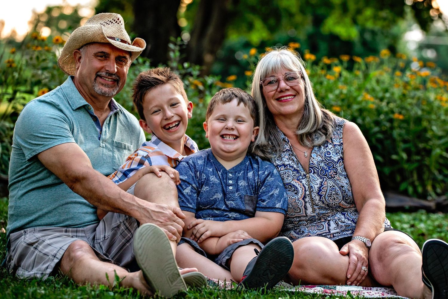 A family of four sits together on grass in a garden setting during a sunny summer day.