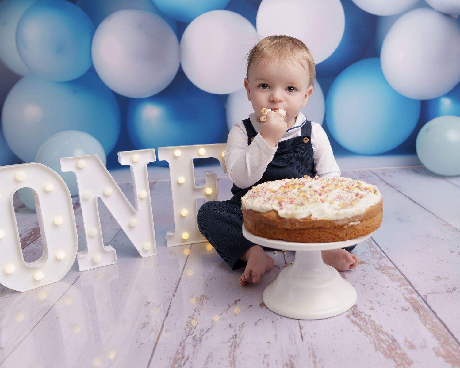 First birthday setup with cake and light-up ONE sign against blue and white balloon backdrop.
