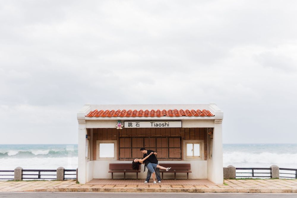 A couple embraces at a seaside bus stop shelter with red terracotta roof tiles and ocean views in the background.