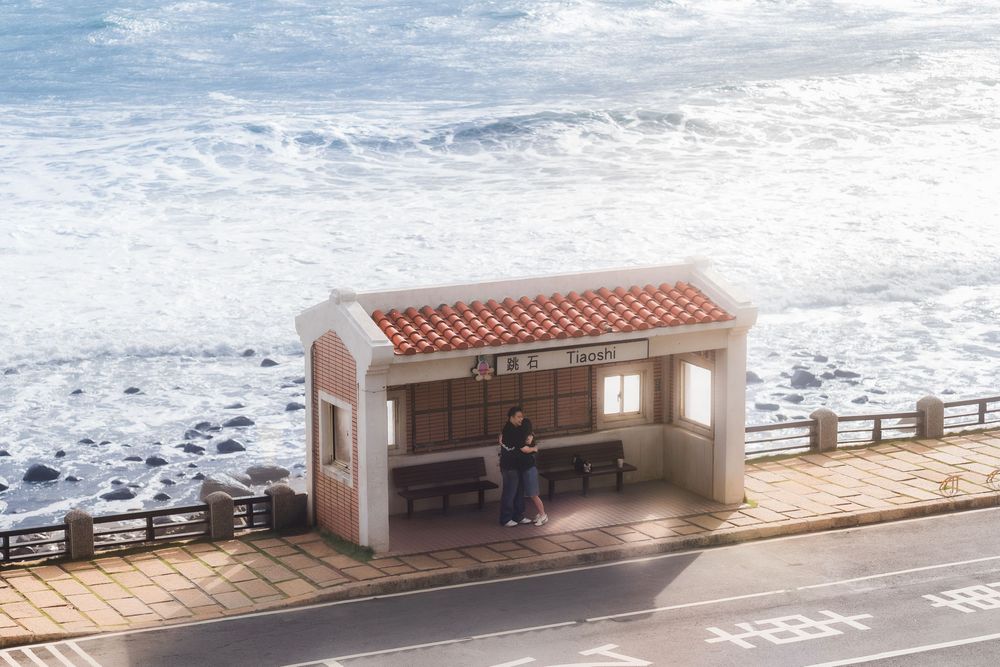 A small beige stucco bus shelter stands along a coastal road with crashing ocean waves in the background.
