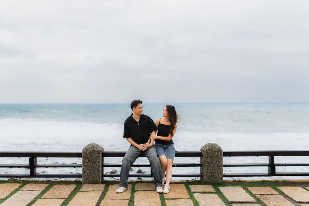 Two people sitting on a wooden bench by the ocean with a cloudy sky in the background.