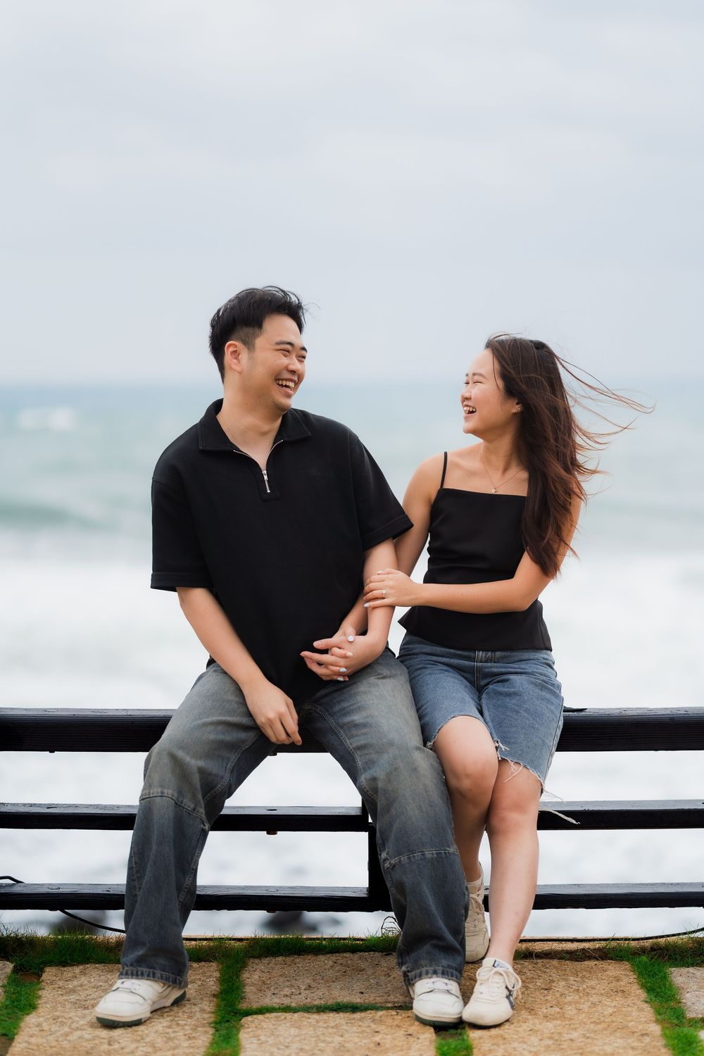 A couple laughs together while sitting on a wooden railing by the seaside.
