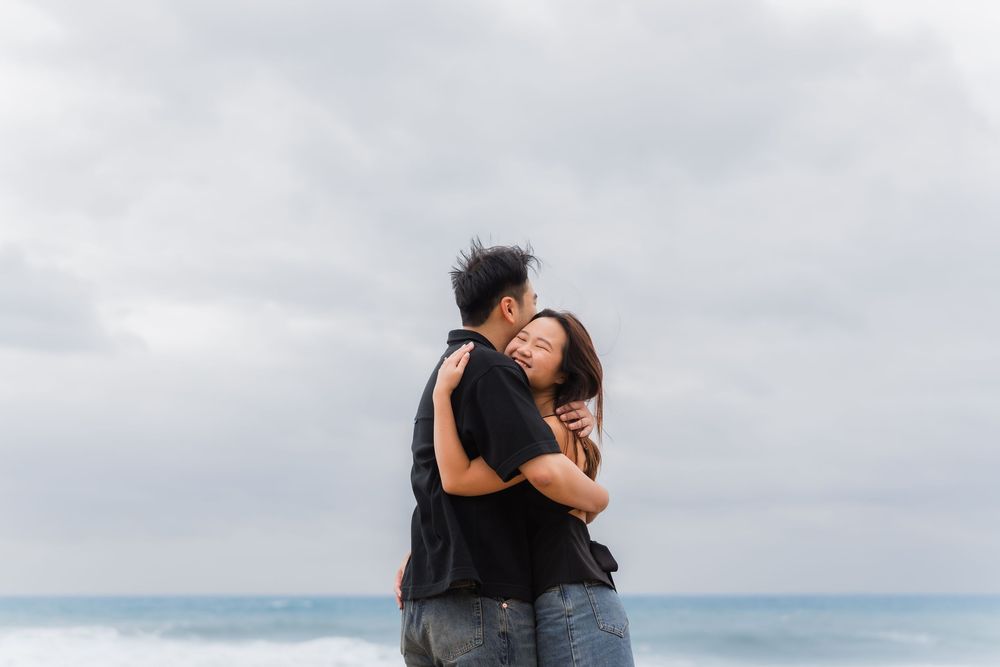 Two people embrace on a beach with cloudy skies and ocean waves in the background.