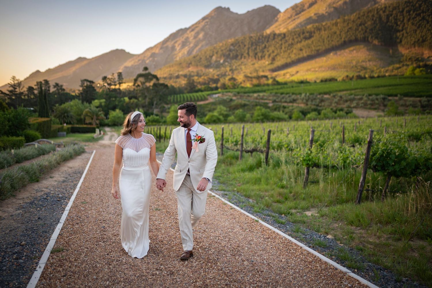 A couple in wedding attire walks hand in hand along a scenic mountain path at sunset.