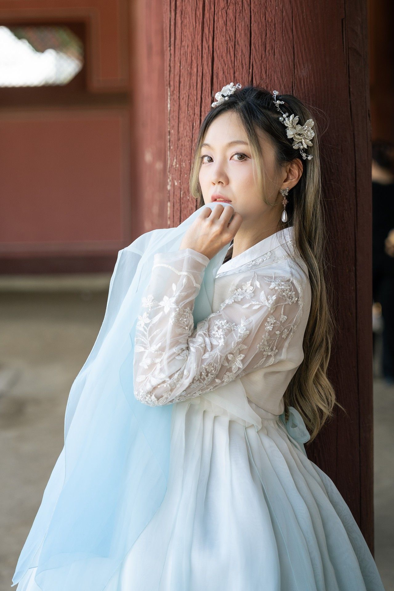 Person in traditional hanbok-inspired white lace dress with floral hair accessories poses thoughtfully by wooden pillar.